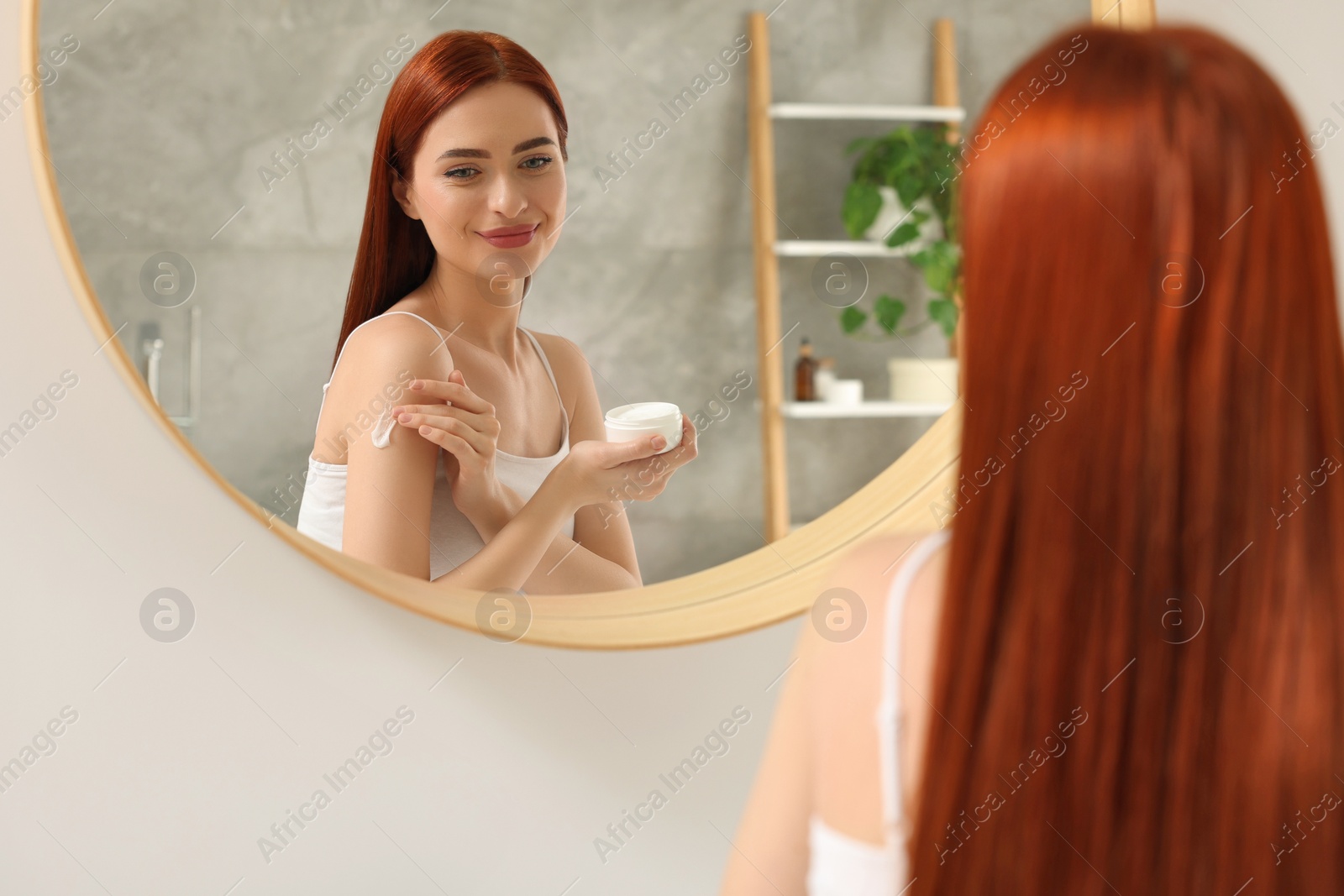 Photo of Beautiful young woman applying body cream onto shoulder near mirror indoors