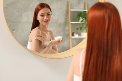 Beautiful young woman applying body cream onto shoulder near mirror indoors