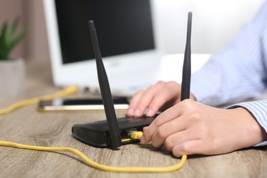 Photo of Woman inserting ethernet cable into Wi-Fi router at wooden table indoors, closeup