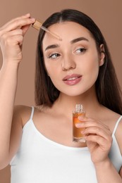 Photo of Beautiful woman applying essential oil onto face on brown background