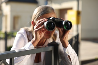 Photo of Concept of private life. Curious senior woman with binoculars spying on neighbours over fence outdoors