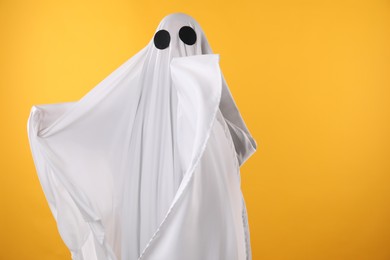 Photo of Creepy ghost. Person covered with white sheet on yellow background, space for text