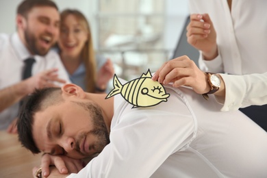 Photo of Young woman sticking paper fish to colleague's back while he sleeping in office, closeup. Funny joke