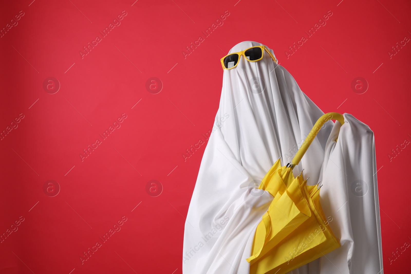 Photo of Person in ghost costume and sunglasses holding yellow umbrella on red background, space for text