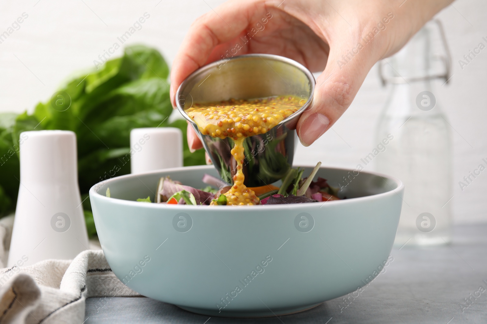 Photo of Woman pouring tasty vinegar based sauce (Vinaigrette) into bowl with salad at grey table, closeup