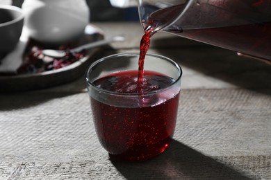 Pouring aromatic hibiscus tea from teapot into glass at wooden table, closeup