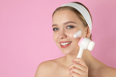 Young woman washing face with brush and cleansing foam on pink background. Cosmetic products