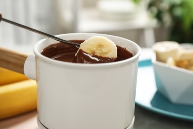 Dipping slice of banana into fondue pot with milk chocolate on table indoors, closeup