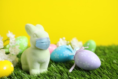 Image of COVID-19 pandemic. Easter bunny toy in protective mask and dyed eggs on green grass