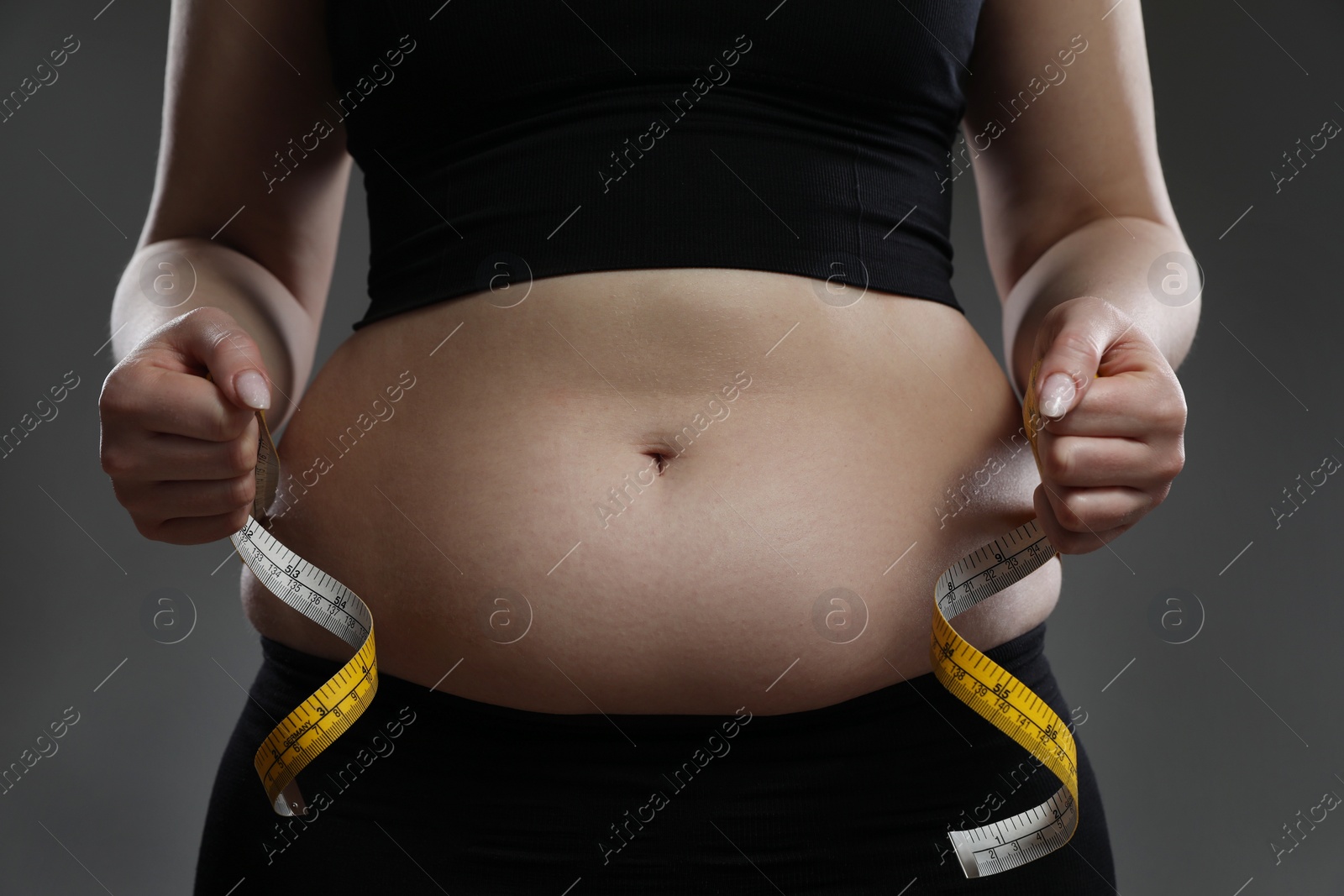 Photo of Woman measuring belly with tape on grey background, closeup. Overweight problem