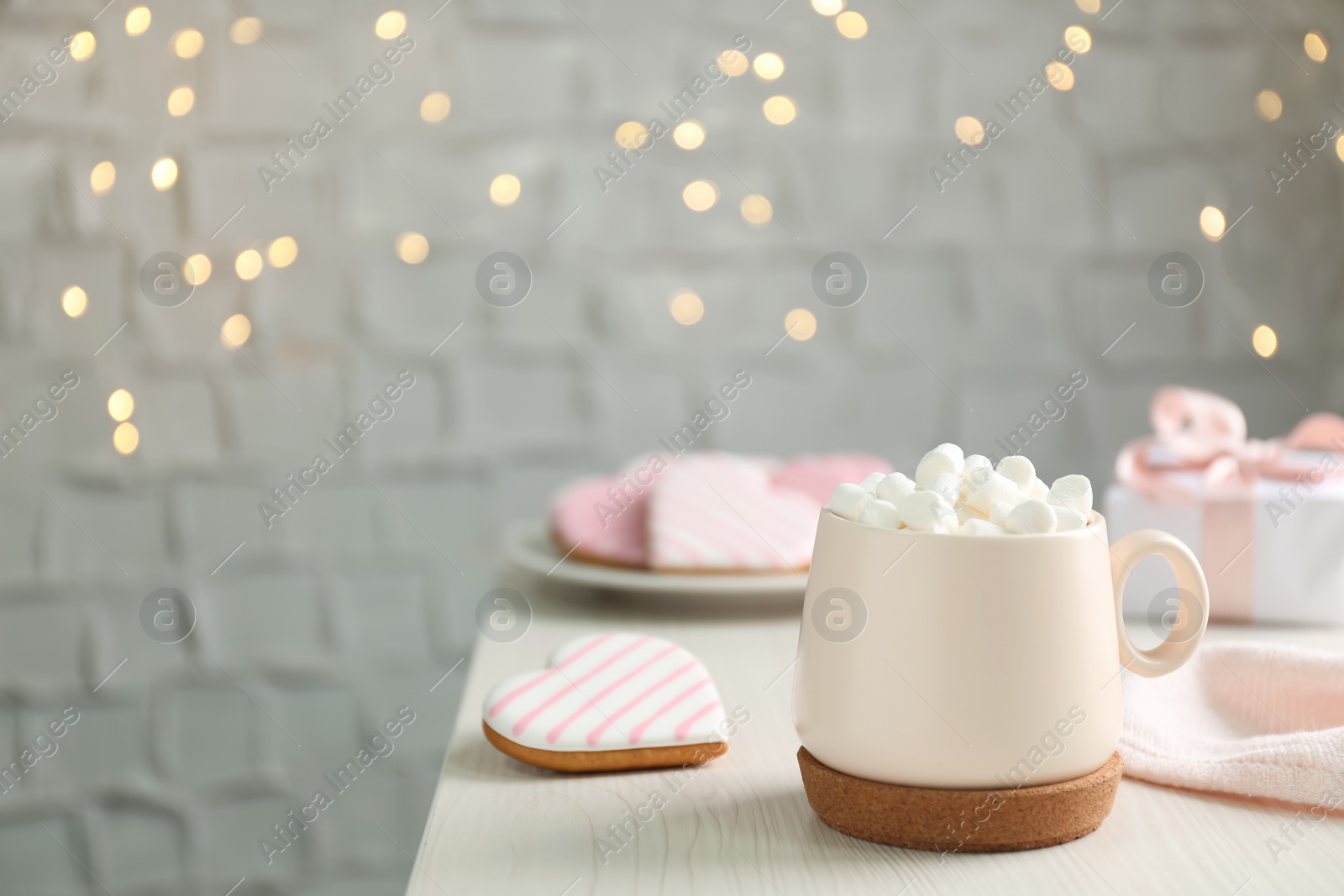 Photo of Cup of delicious drink with marshmallows and heart shaped cookie on white table against blurred lights, space for text