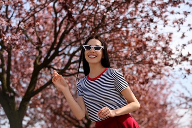 Pretty young woman with sunglasses near beautiful blossoming trees outdoors. Stylish spring look