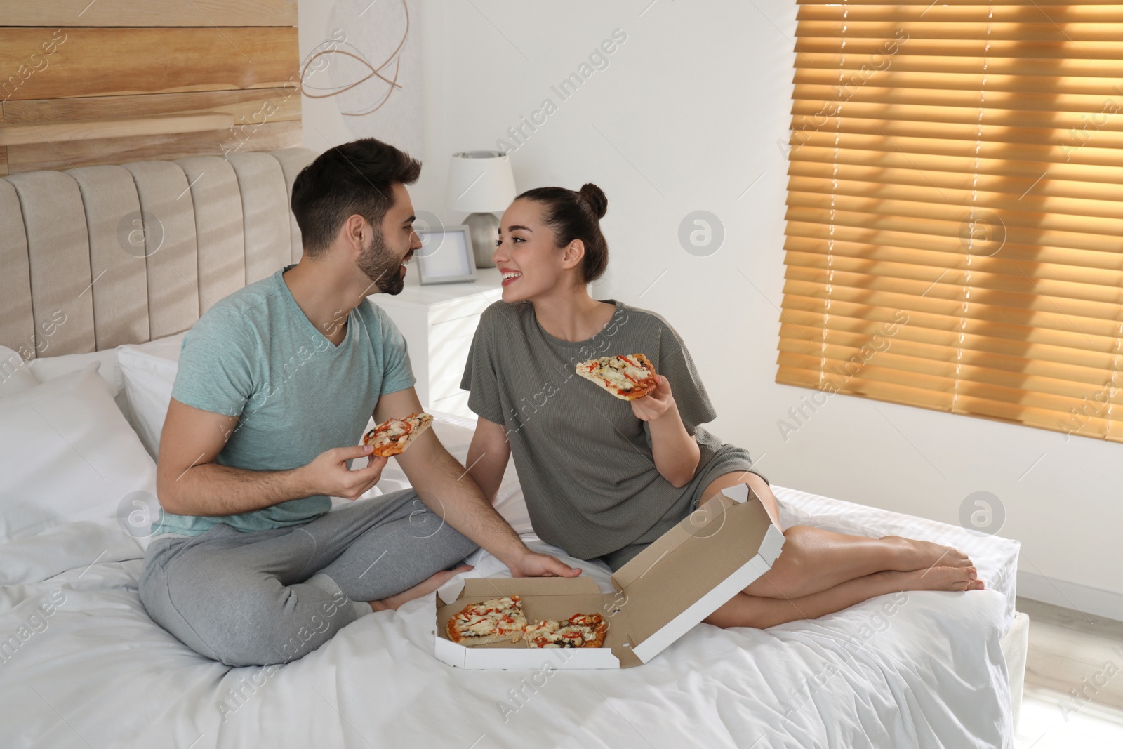 Photo of Happy couple in pajamas eating pizza on bed at home