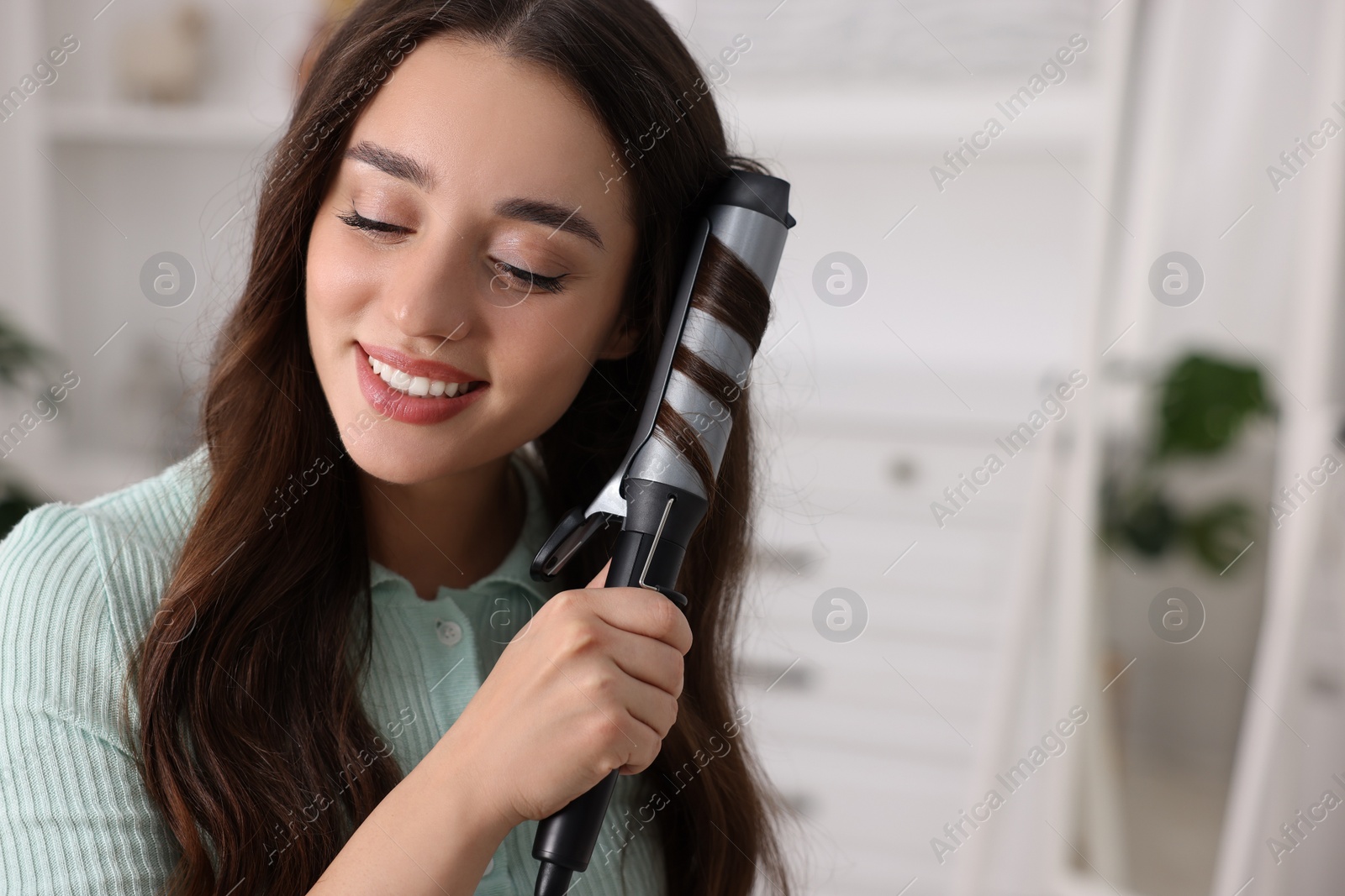 Photo of Smiling woman using curling hair iron at home. Space for text