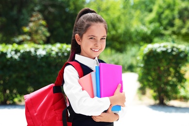 Photo of Cute little girl in school uniform with backpack and stationery on street