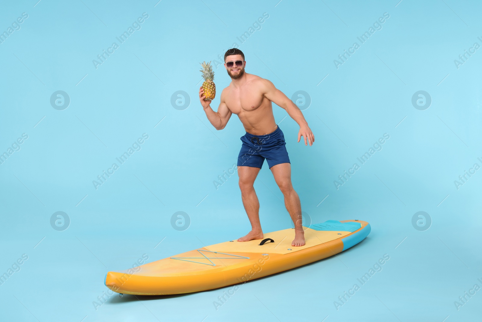 Photo of Happy man with pineapple posing on SUP board against light blue background