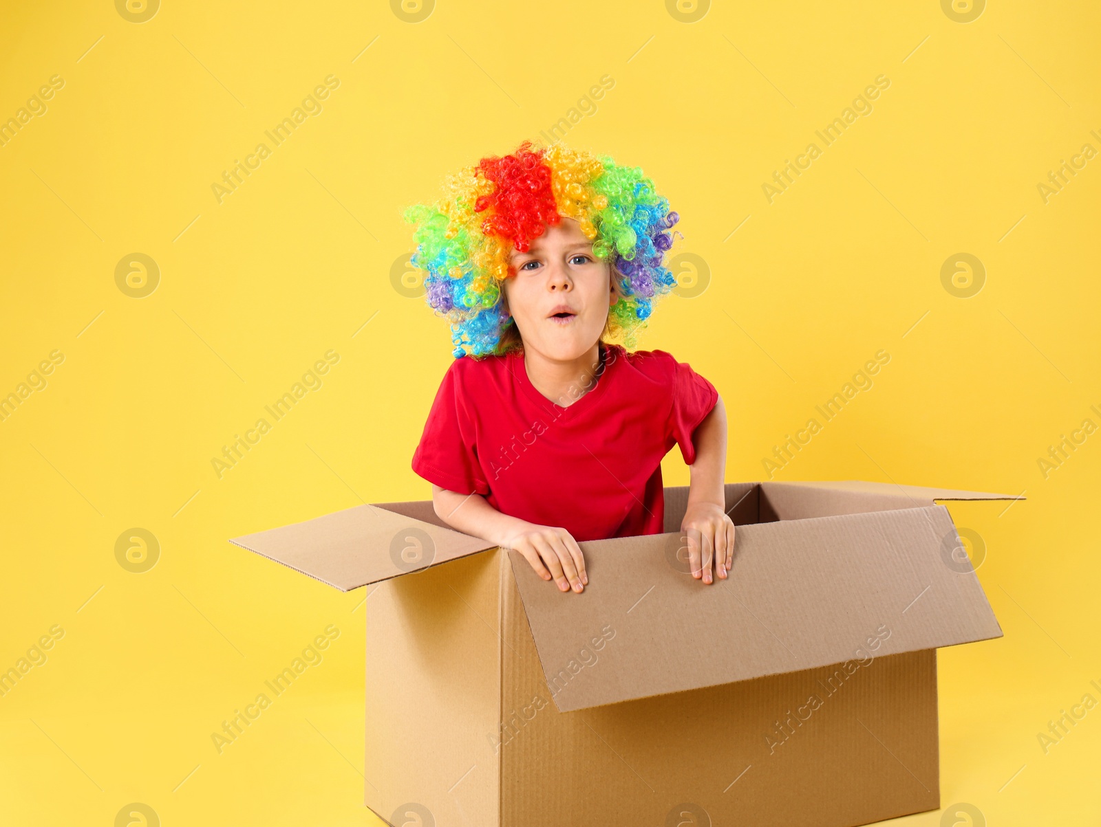 Photo of Funny little boy in clown wig getting out of cardboard box on yellow background. April fool's day