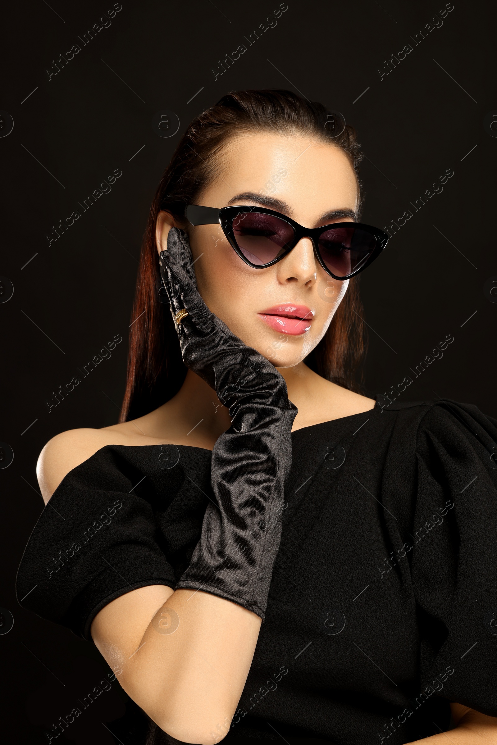 Photo of Portrait of beautiful young woman in sunglasses and elegant evening gloves on black background