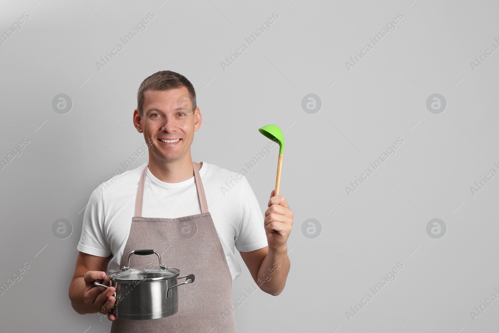 Photo of Happy man with cooking pot and ladle on light grey background. Space for text