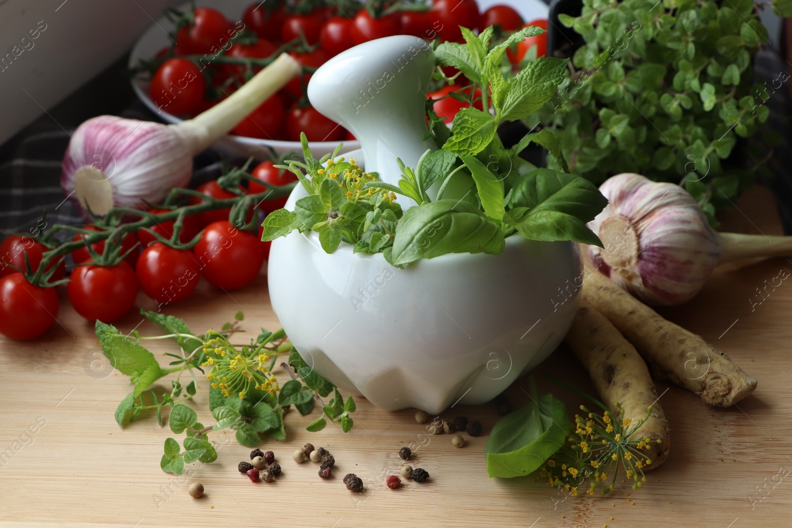 Photo of Mortar with fresh herbs near garlic, horseradish roots, black peppercorns and cherry tomatoes on wooden table, closeup