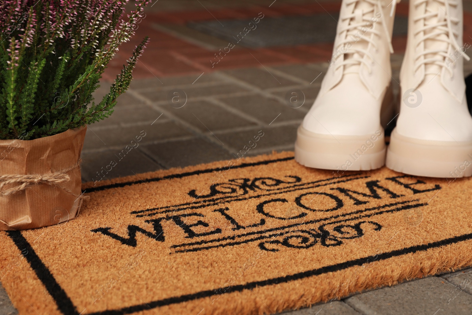 Photo of Door mat with word Welcome, stylish boots and beautiful flowers on floor, closeup