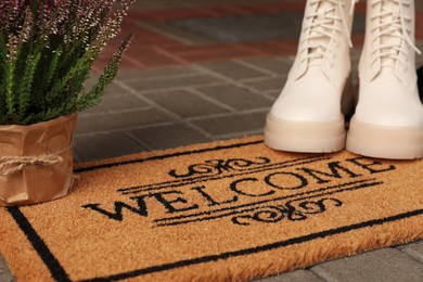 Photo of Door mat with word Welcome, stylish boots and beautiful flowers on floor, closeup