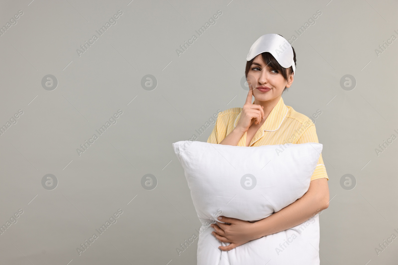 Photo of Woman in pyjama and sleep mask holding pillow on grey background, space for text