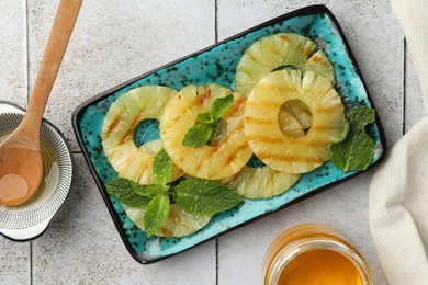 Tasty grilled pineapple slices, honey and wooden spoon on light gray tiled table, flat lay