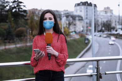 Image of Young journalist with medical mask, microphone and smartphone working on city street. Virus protection
