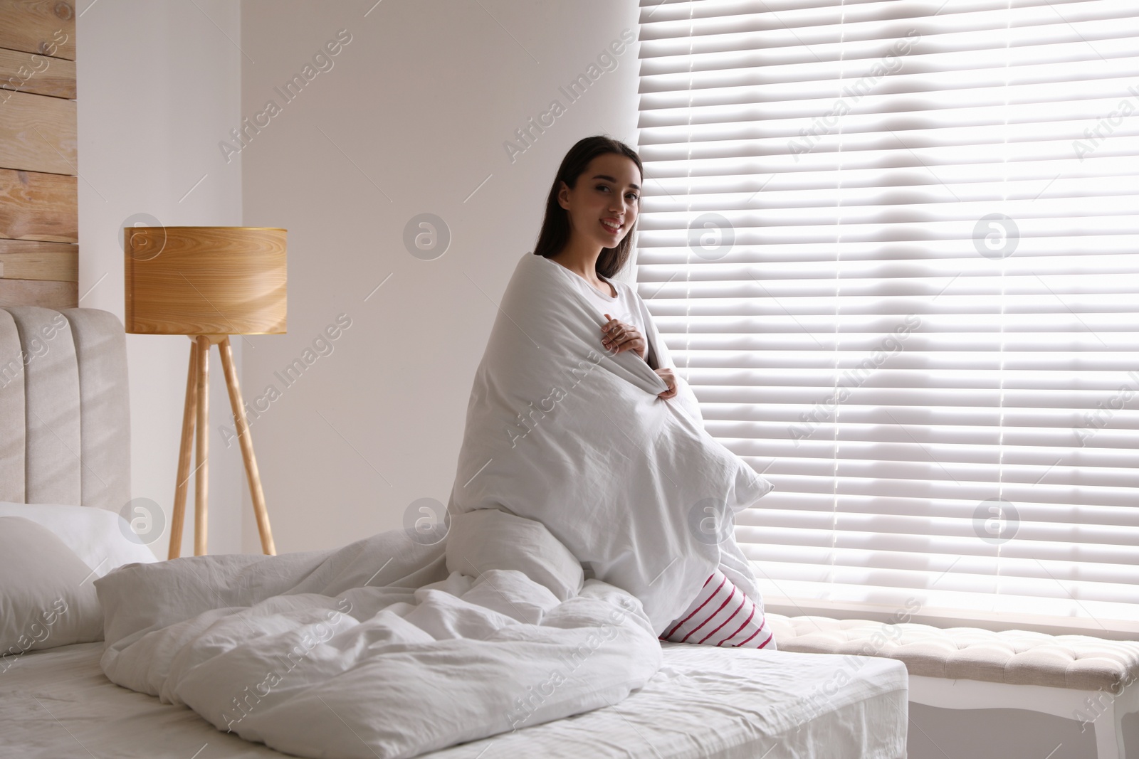 Photo of Beautiful young woman covered with warm blanket near window at home