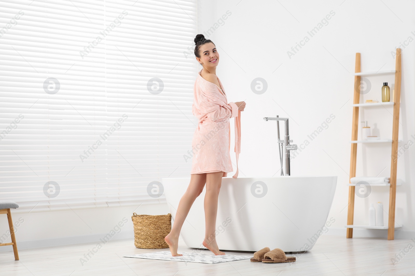 Photo of Beautiful happy woman wearing stylish bathrobe near tub in bathroom