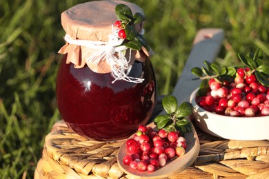 Photo of Jar of delicious lingonberry jam and red berries on wicker basket outdoors