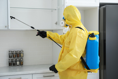 Photo of Pest control worker in protective suit spraying insecticide on furniture indoors