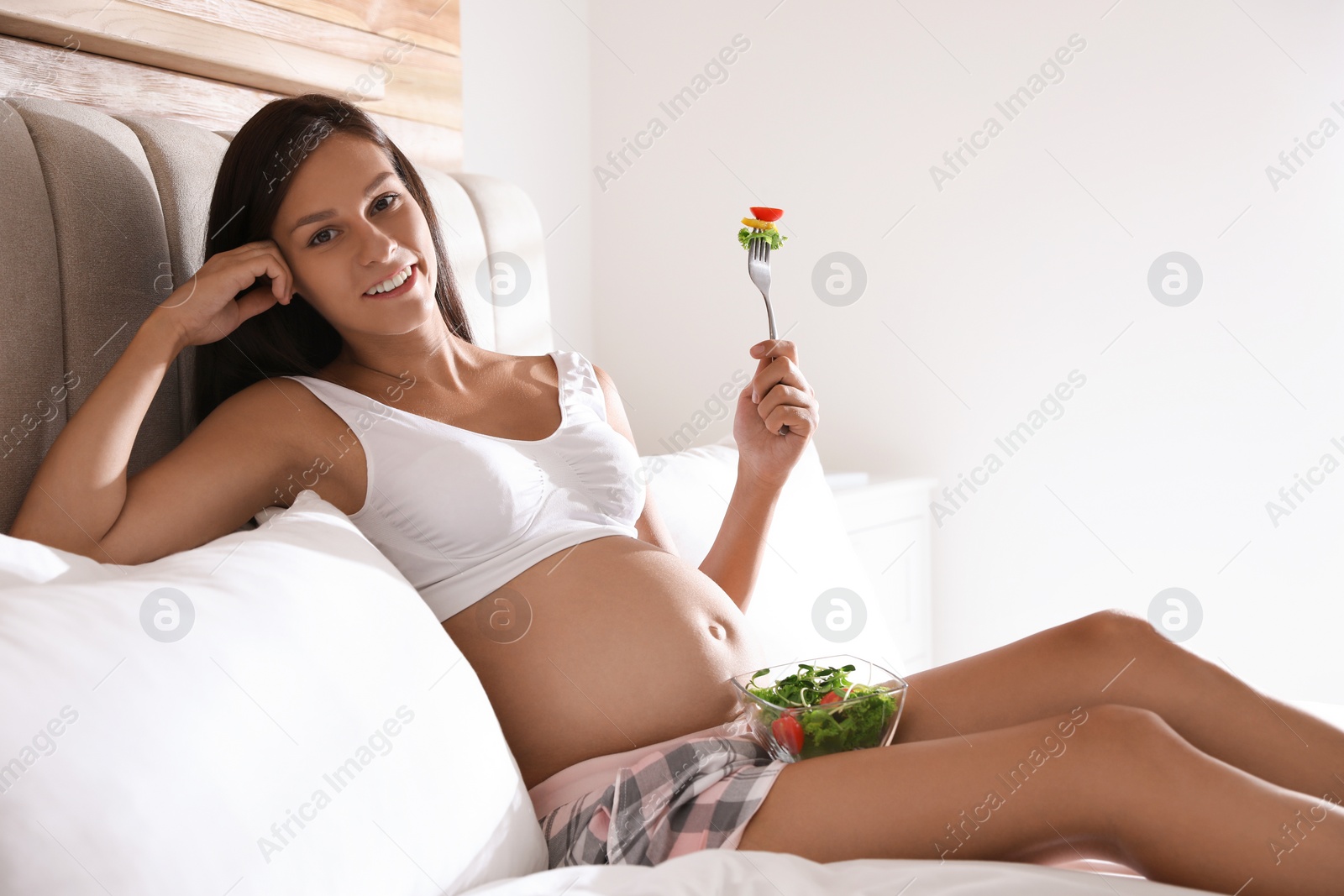 Photo of Young pregnant woman with bowl of vegetable salad in bedroom. Taking care of baby health