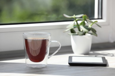 Cup of tea, e-book reader and houseplant on wooden window sill