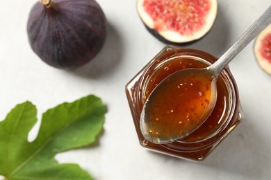 Photo of Glass jar of tasty sweet jam with spoon, green leaf and fresh figs on white table, flat lay