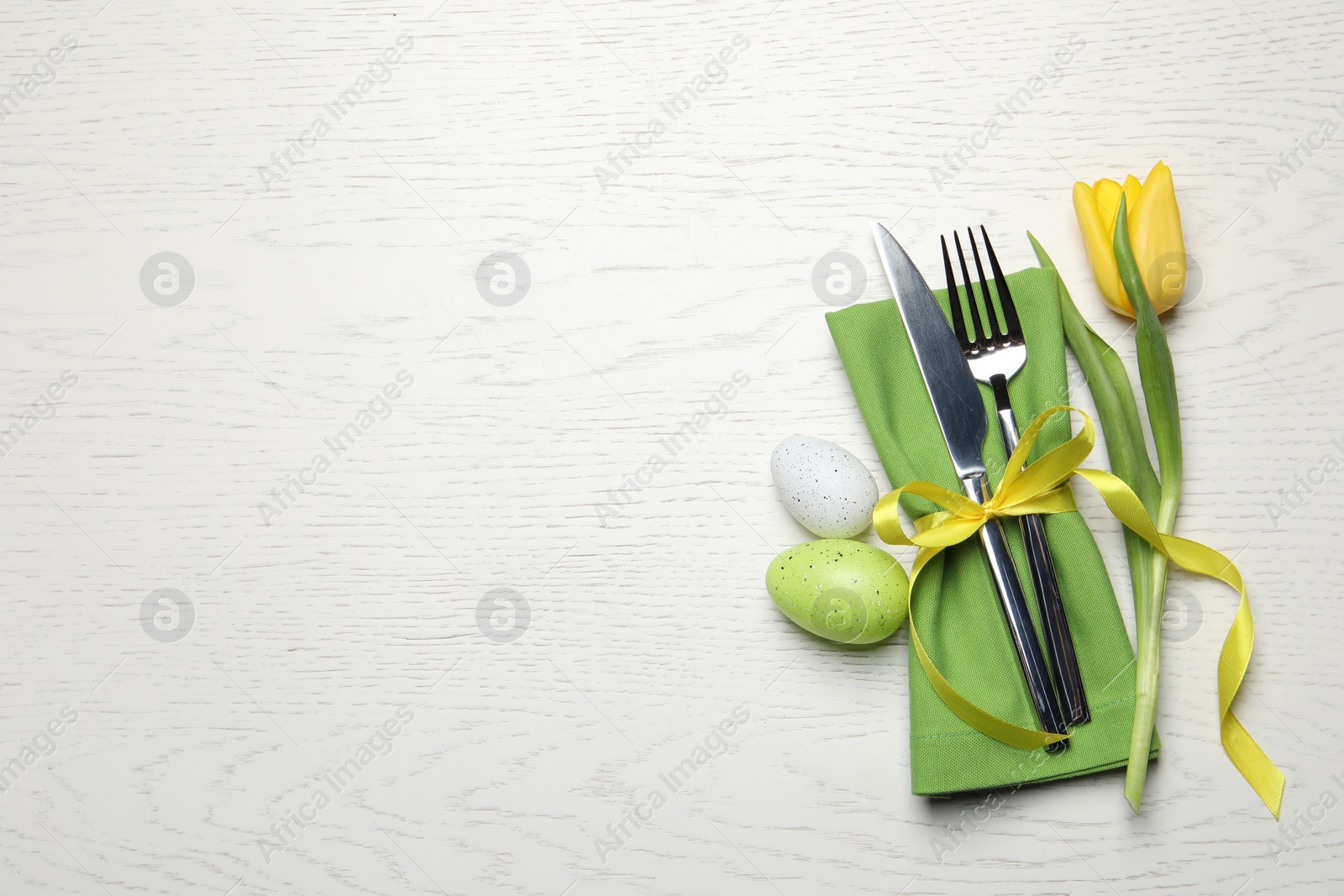 Photo of Cutlery set, painted eggs and beautiful flower on white wooden table, flat lay with space for text. Easter celebration