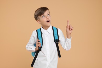 Surprised schoolboy with backpack pointing upwards on beige background