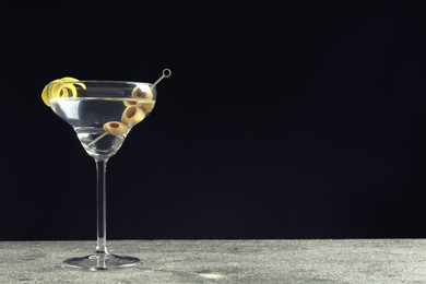 Photo of Martini cocktail with olives and lemon twist on grey table against dark background, space for text