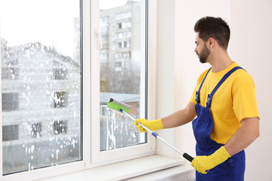 Professional young janitor in uniform cleaning window indoors