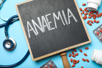 Small blackboard with word Anaemia, pills and stethoscope on light blue background, flat lay