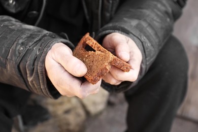 Photo of Poor homeless man holding piece of bread outdoors, closeup