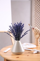 Photo of Bouquet of beautiful preserved lavender flowers, palette, paints and manikin hand with brush on wooden table near easel indoors, closeup