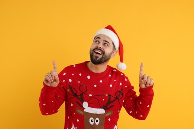Photo of Happy young man in Christmas sweater and Santa hat pointing at something on orange background
