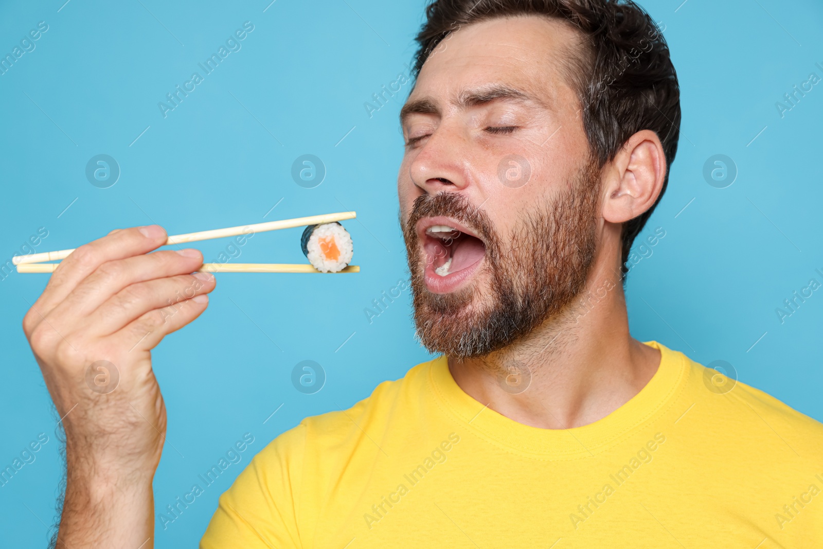 Photo of Handsome man eating sushi roll with chopsticks on light blue background