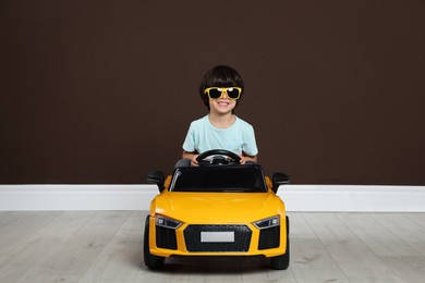 Cute little boy driving children's electric toy car near brown wall indoors