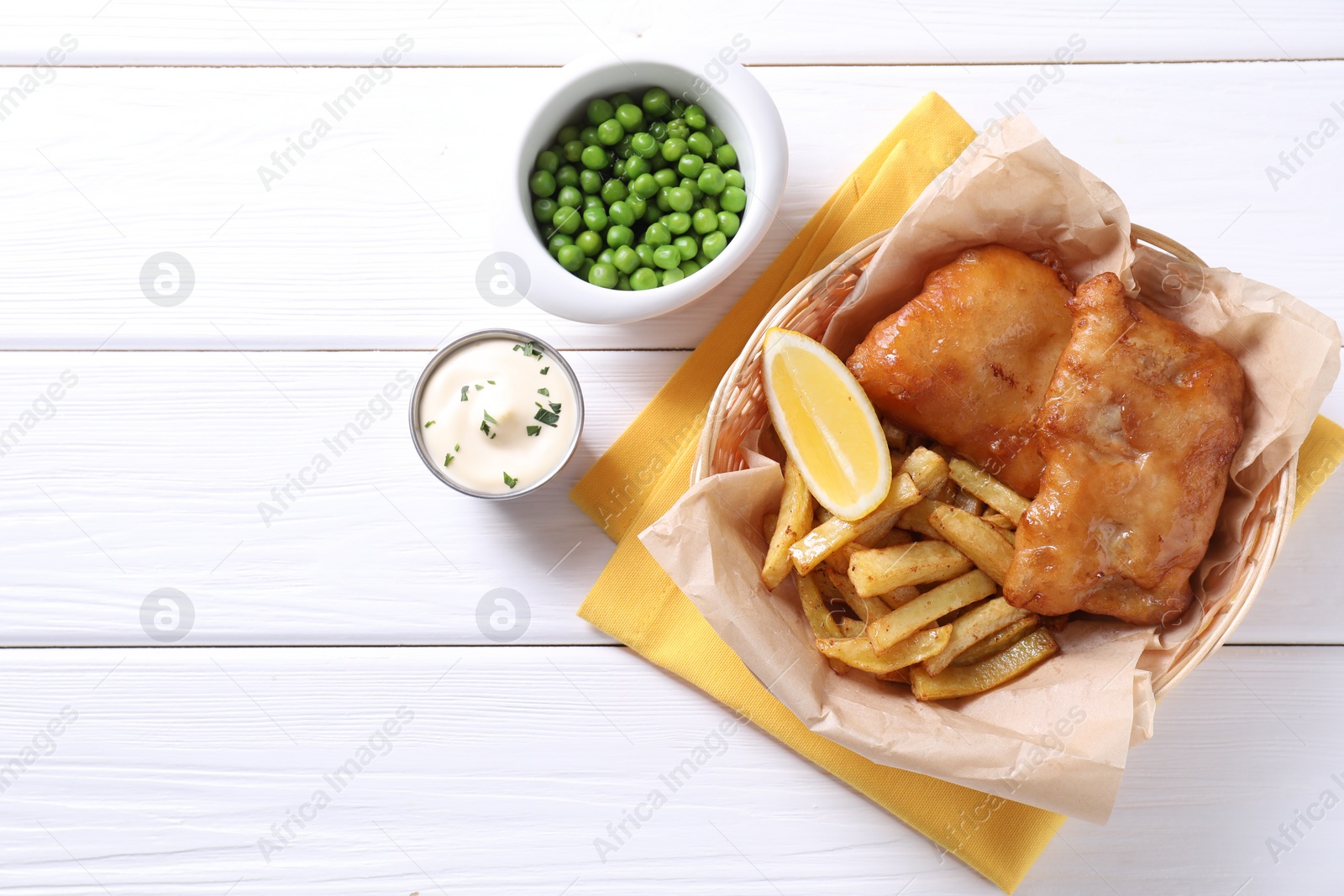 Photo of Tasty fish, chips, sauce, peas and lemon in wicker bowl on white wooden table, top view. Space for text