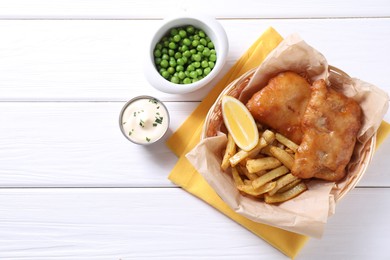 Tasty fish, chips, sauce, peas and lemon in wicker bowl on white wooden table, top view. Space for text