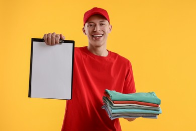 Photo of Dry-cleaning delivery. Happy courier holding folded clothes and clipboard on orange background