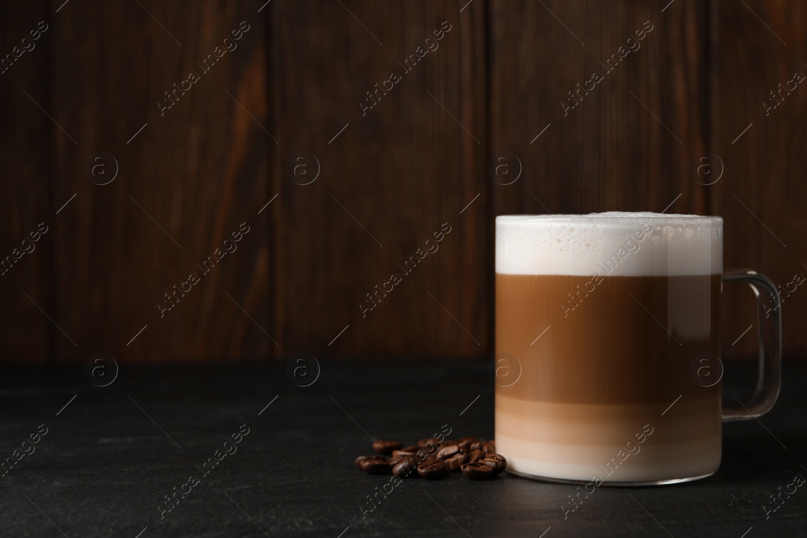 Photo of Glass cup of delicious layered coffee and beans on black table against wooden background, space for text